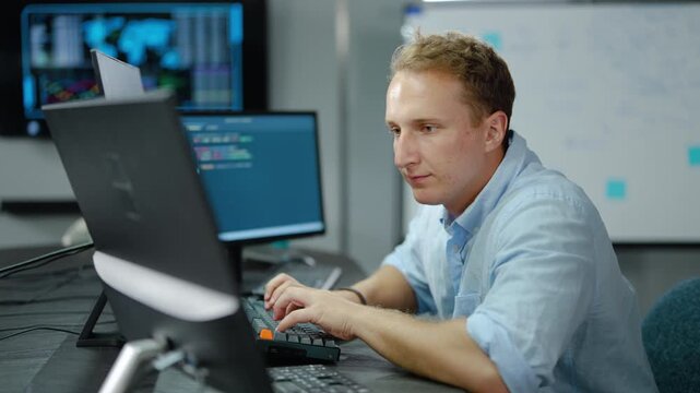 Focused male software developer typing on a computer keyboard at a desk with multiple monitors displaying complex code and data analytics in a modern, dark high-tech corporate office environment.