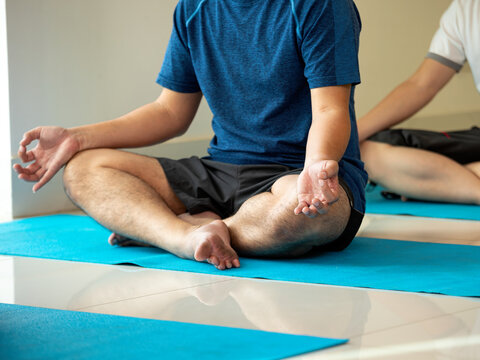 Man practicing yoga meditation in lotus pose on a blue mat indoors, showing mindfulness, wellness, balance, relaxation, breathing, fitness, focus, and calm group exercise