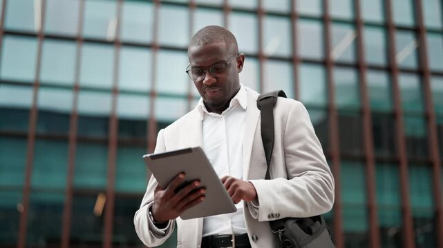 Black businessman, using tablet, browsing report, outdoor near office building. African american business man smiling, works as marketing director, staying connected with technology, leading projects.