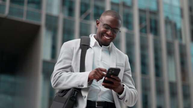 African american businessman, using smartphone, texting, outdoor near office building. Black business man smiling, works as marketing manager, focused on daily tasks, driving quarterly growth targets.