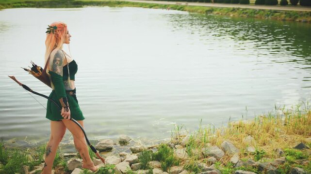 Fantasy elf woman walking along lake shore clear blue water pond. happy fairy girl princess archer beauty face fashion model posing. lady enjoys wild nature green grass bright sun light river bank