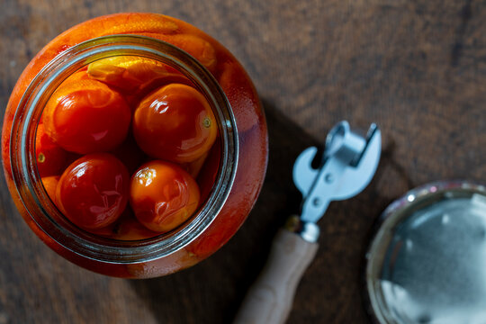 Open jar of pickled cherry tomatoes on wooden background, closeup. Top view of open glass jar filled with marinated orange cherry tomatoes next vintage can opener