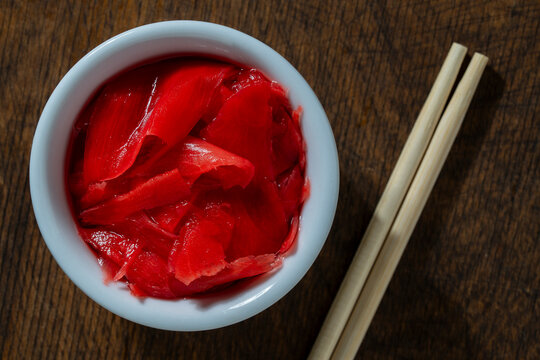 Pickled pink ginger in bowl with chopsticks, closeup. Top view of white ceramic bowl filled with red pickled sushi ginger next to wooden chopsticks on dark background