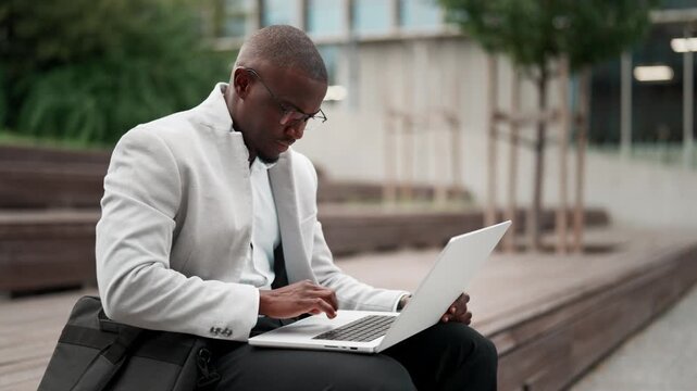 Black businessman, sitting, using laptop, typing report, outdoor near office building. African american business man smiling, works as operations manager, staying connected with technology. Copy space