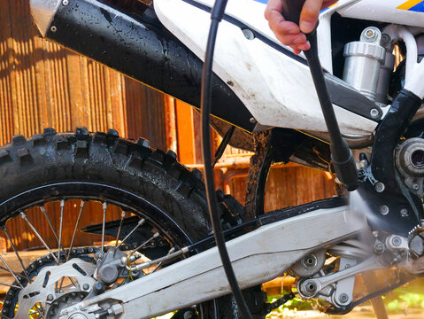 Close-up of a person bending over to hold a pressure washer gun and clean the rear wheel area of a white dirt bike outdoors.