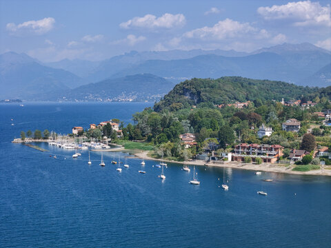 Aerial view of Sasso Moro headland on Lake Maggiore with sailboats moored in the bay, surrounded by lush greenery and villas, with distant mountains in Leggiuno, Lombardia, Italy.