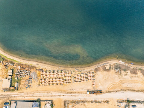 Aerial view of Kali Baru showing rows of stacked wooden materials on the sandy shore next to the calm sea, with nearby industrial structures and a coastal road in Cilincing, Jakarta, Indonesia.