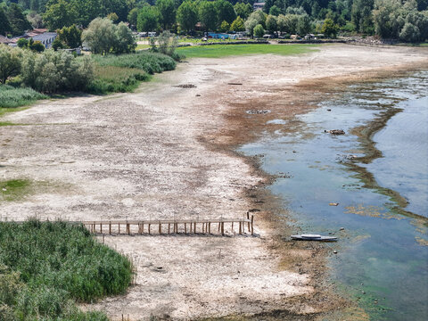 Aerial view of the receding shoreline of Lake Maggiore during a severe drought, revealing exposed dry lakebed, a stranded wooden pier, and a small boat in Monvalle, Lombardy, Italy.