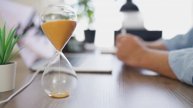 Businessman turning over an hourglass while sit at office desk, preparing to begin an important task. Time management, productivity, planning and workplace efficiency concept