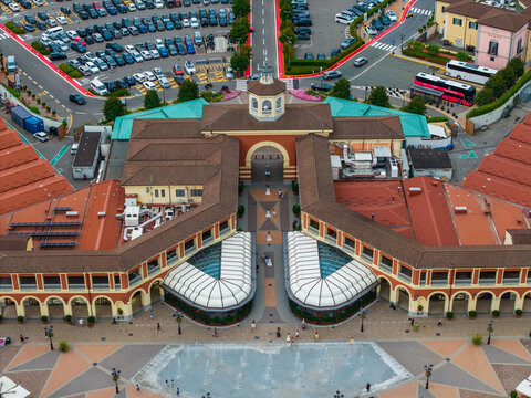 Aerial view of Designer Outlet Serravalle featuring a central fountain plaza, terracotta-roofed retail buildings, and surrounding parking areas in Serravalle Scrivia, Piemonte, Italy.