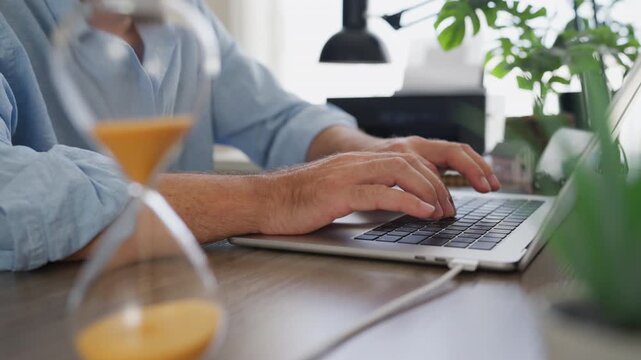 Businessman turning over an hourglass while working on laptop at office desk. Time management, productivity, deadline, multitasking, workflow and business efficiency concept.