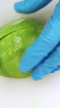 Ripe avocado cutting close up on white kitchen board. Chef hands in blue gloves mince fresh vegetable with big iron knife. Cooking, preparing ingredients in professional restaurant. Top down view