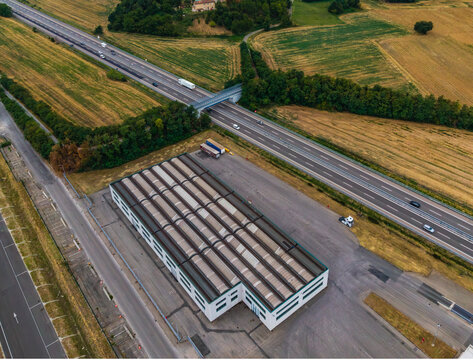 Aerial view of the Roquette plant facility with industrial buildings and corrugated roofs, near a highway with vehicles, surrounded by fields in Serravalle Scrivia, Piedmont, Italy.