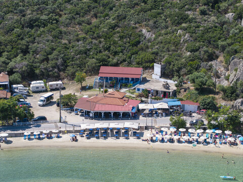 Aerial view of Sykias Beach with a sandy shoreline, umbrellas, sun loungers, a restaurant with a red-tiled roof, parked caravans, and a lush green hillside in Paralia Sikias, Greece.