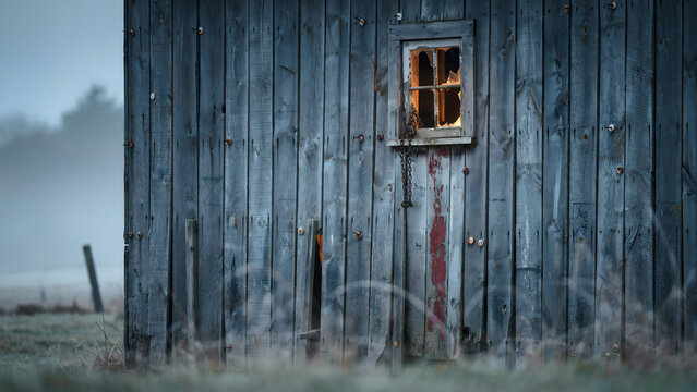 A close-up view of a weathered blue wooden barn wall with a small, broken window pane glowing with warm light against a foggy, overcast sky