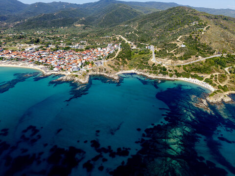 Aerial view of Sarti Beach with its turquoise waters, rocky coastline, and the town of Sarti featuring red-roofed houses nestled against lush green hills in Greece.