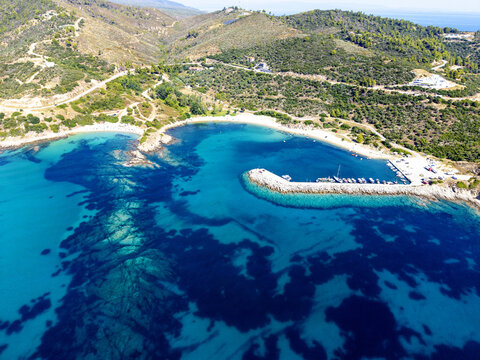 Aerial view of Sarti Beach featuring a small harbor with moored boats, turquoise waters, and rugged hills in Greece.