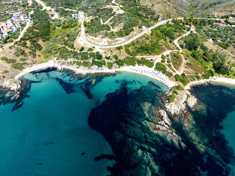 Aerial view of Sarti Beach featuring turquoise waters, rocky coastal formations, a sandy shoreline with umbrellas, and a winding road along the hillside in Greece.