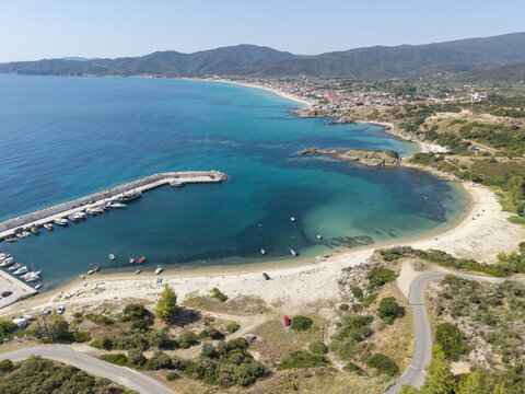 Aerial view of Sarti Beach featuring turquoise waters, a sandy shoreline, a small harbor with docked boats, and the coastal town in Sarti, Greece.