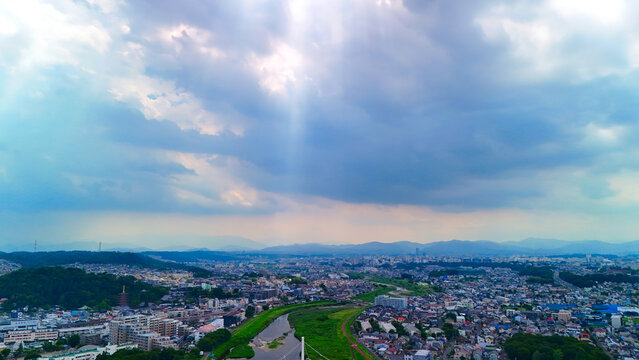 Aerial view of the Asakawa river winding through the urban landscape, with green banks, residential buildings, and distant mountains under a dramatic cloudy sky Hino, Tokyo, Japan.