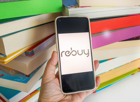 Braunschweig, Germany - August 06, 2026: Hand holding smartphone with Rebuy logo on screen against stack colorful books, sustainable recommerce, circular economy, and buying used textbooks online.