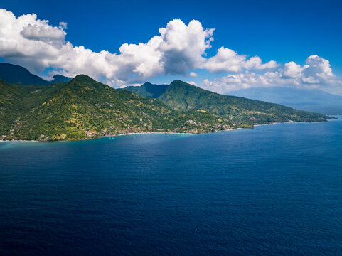 Aerial view of the Amed coastline featuring lush green mountains meeting the deep blue sea under a bright sky with fluffy white clouds in Abang, Bali, Indonesia.