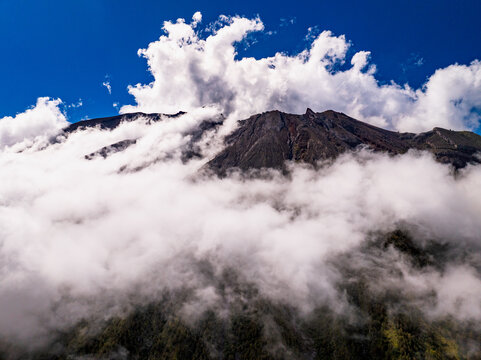Aerial view of Mount Agung with volcanic slopes partially covered by thick white clouds under a clear blue sky in Bebandem, Bali, Indonesia.
