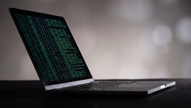 Laptop screen displays lines of glowing green computer code against a blurred and moody background.