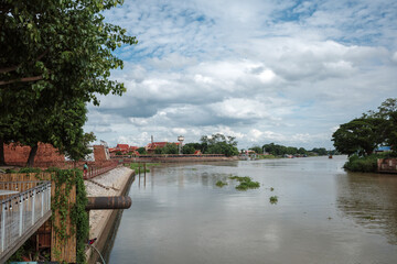 Fototapeta premium A scenic exterior view of Phra Nakhon Fortress located between the Chao Phraya River and the Pasak River in Ayutthaya, Thailand, featuring historic brick walls, a wooden waterfront structure, trees, a