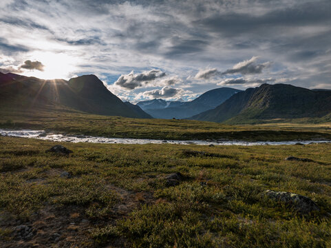 Aerial view of Jotunheimen National Park with a winding river flowing through rugged mountains under a dramatic cloudy sky with sun flare in Lom Municipality, Innlandet, Norway.