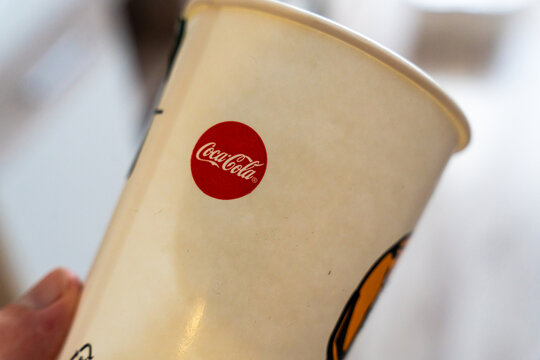 Warsaw, Poland - July 30, 2026: Coca-Cola branded paper cup held by hand in close-up, showing red circular logo on beige disposable drink container