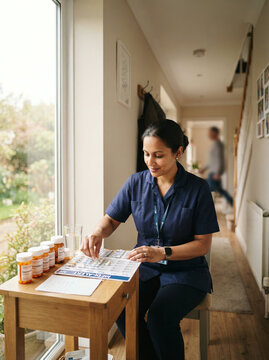 Healthcare worker on a stool by a table of pill bottles in a bright hallway