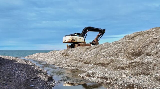 Excavator machine sits atop pebble mound near black sea coastline during beach construction work.