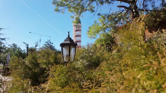 Decorative vintage street lamp stands among dense green bushes with tower and blue sky.
