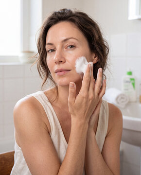 Woman in bathroom applying white cleansing foam to her cheek in a natural morning routine