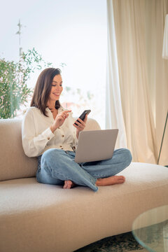 Attractive woman sitting comfortable on the couch and smiling while using mobile phone and laptop, home office