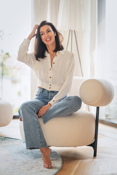 Full length of a cheerful smiling woman sitting in an armchair in her modern home