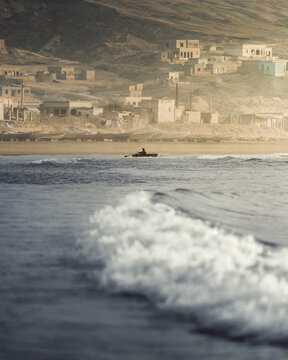 Aerial view of a lone fisherman rowing a small wooden boat on calm coastal waters in front of a traditional village with stone houses nestled against arid hills.