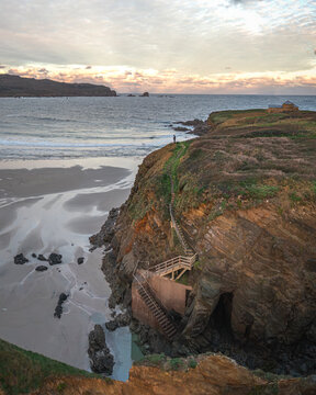 Aerial view of Ermita de Santa Comba chapel on a rocky cliff overlooking a sandy beach with a wooden staircase and a person at sunset Covas, La Coruna, Spain.