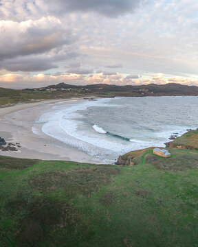 Aerial view of Ermita de Santa Comba on a green cliff overlooking a sandy beach with crashing waves and rolling hills under a cloudy sky in Covas, La Coruna, Spain.