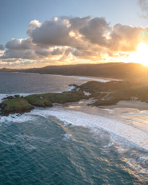 Aerial view of Ermita de Santa Comba on a rocky islet surrounded by crashing waves and a sandy beach under a golden sunset Covas, La Coruna, Spain.
