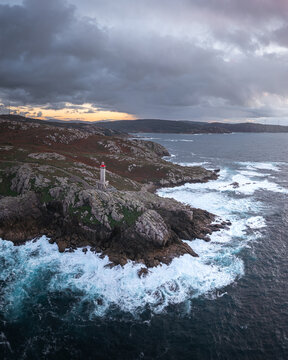Aerial view of the Faro de Punta Nariga lighthouse standing on rugged rocky cliffs with crashing waves under a dramatic cloudy sky in Malpica, A Coruna, Spain.