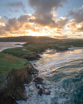 Aerial view of Ermita de Santa Comba on a rocky cliff overlooking the beach and crashing waves during a golden sunset in Covas, La Coruna, Spain.