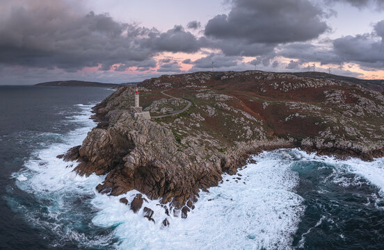 Aerial view of Faro de Punta Nariga lighthouse on rugged rocky cliffs with crashing white waves and wind turbines under a dramatic cloudy sky in Malpica, A Coruna, Spain.