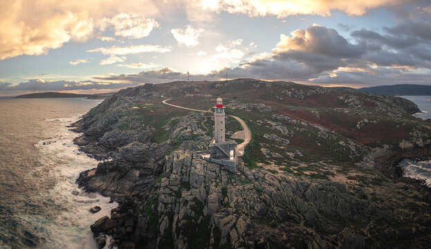 Aerial view of Faro de Punta Nariga lighthouse standing on rugged rocky cliffs overlooking the Atlantic Ocean under a dramatic sunset sky with golden clouds Malpica, A Coruna, Spain.