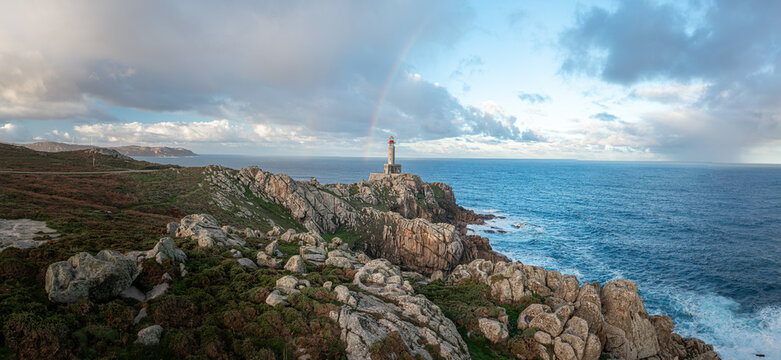 Aerial view of Faro de Punta Nariga lighthouse on a rugged rocky cliff with a rainbow appearing in the cloudy sky over the Atlantic Ocean in Malpica, A Coruna, Spain.