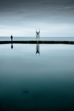12 April 2022: View of a person standing on a concrete pier next to a diving board reflected in the calm water under a cloudy sky.