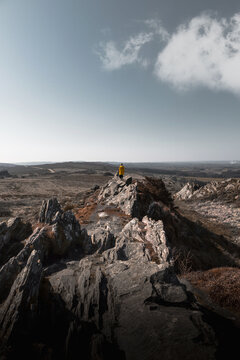 01 March 2021: View of a person in a yellow raincoat standing on a rocky ridge overlooking a vast hilly landscape with dry vegetation under a clear sky with a single cloud.