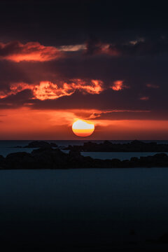 12 April 2022: View of a dramatic sunset over the sea with the sun on the horizon and dark rock silhouettes under a fiery sky.