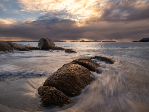 View of large granite boulders on the sandy shore with long exposure waves washing over them under a dramatic sunset sky in Frenchman Bay, Western Australia, Australia.
